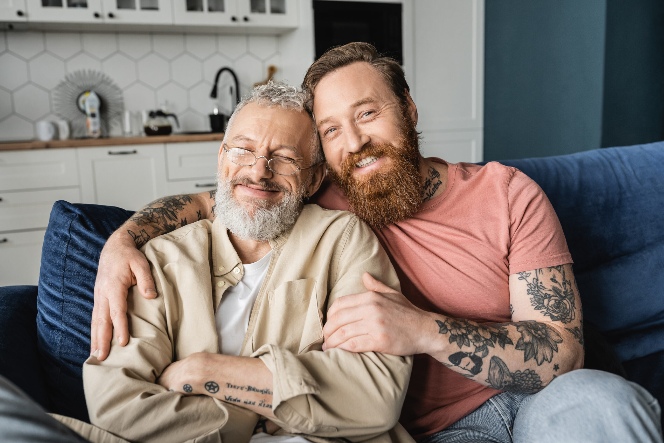 Cheerful gay man hugging bearded partner while sitting on couch at home
